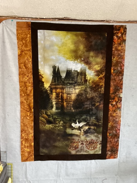 A detailed art piece featuring a large, grand mansion surrounded by lush trees and a serene body of water with swans. The picture is framed with a decorative border in warm autumn colors.