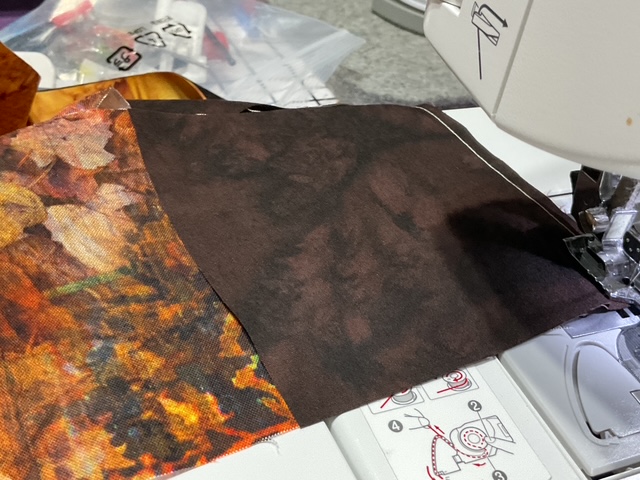 Close-up of a sewing machine with two fabric pieces, one featuring a colorful leaf pattern and the other a solid dark color, being stitched together.