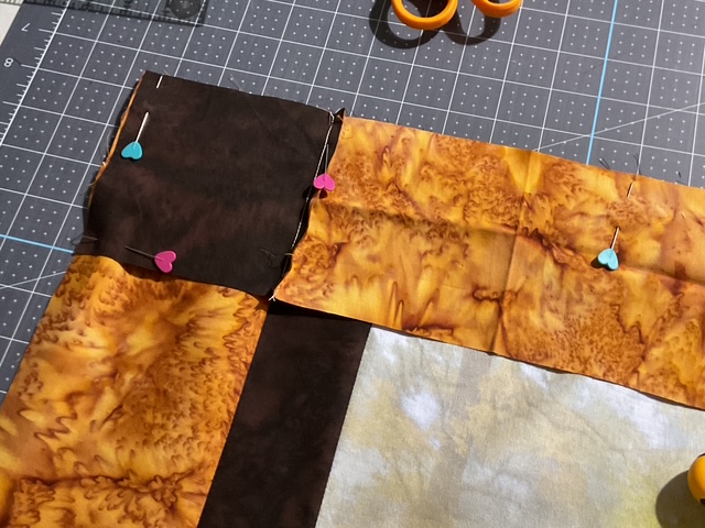 A close-up view of fabric pieces pinned together with colorful heart-shaped pins on a cutting mat, with scissors visible in the background.