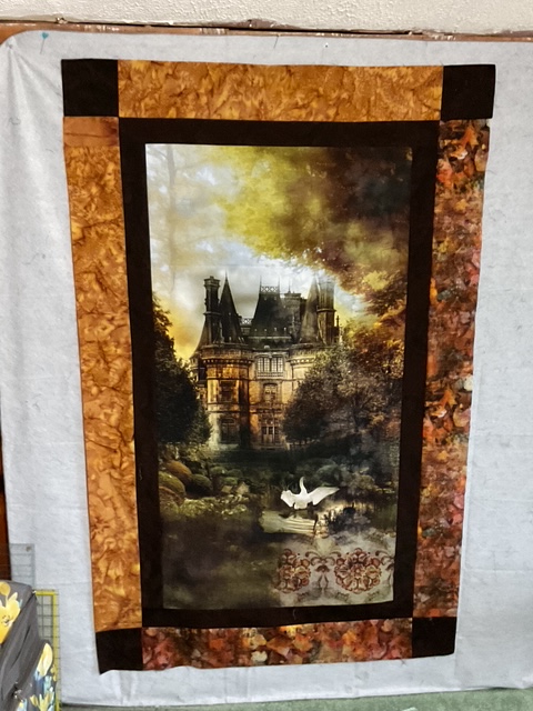 A decorative quilt featuring a printed image of a majestic mansion surrounded by lush trees and a serene pond with swans, framed by a textured brown border.