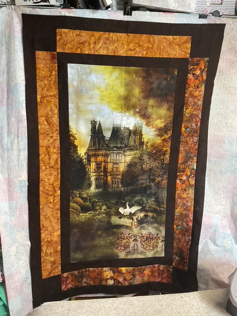 A decorative quilt featuring a printed image of a grand, historic mansion surrounded by lush gardens and trees, framed by fabric in warm colors.