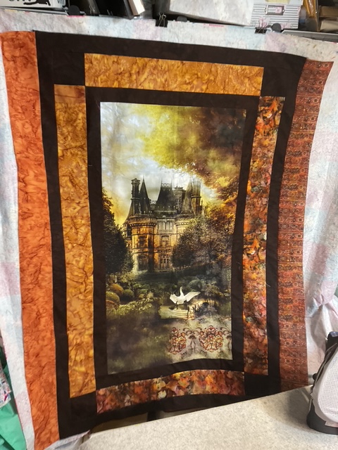 A quilt featuring a central image of a majestic mansion surrounded by trees and a serene landscape, bordered by colorful fabric in autumn hues.