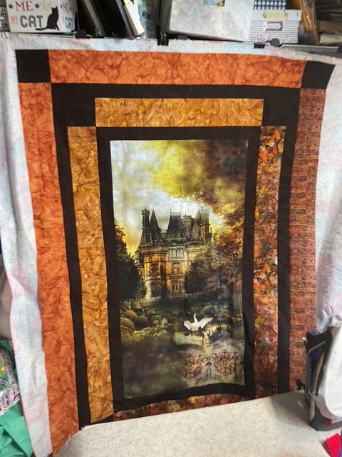 A quilt featuring an image of a grand mansion surrounded by trees and water, with swans in the foreground, bordered by vibrant orange and brown fabric.