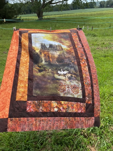 A colorful quilt hanging on a clothesline in a grassy field. The quilt features a central image of a detailed house surrounded by a scenic landscape with trees and a lake, bordered by warm-toned fabric panels.