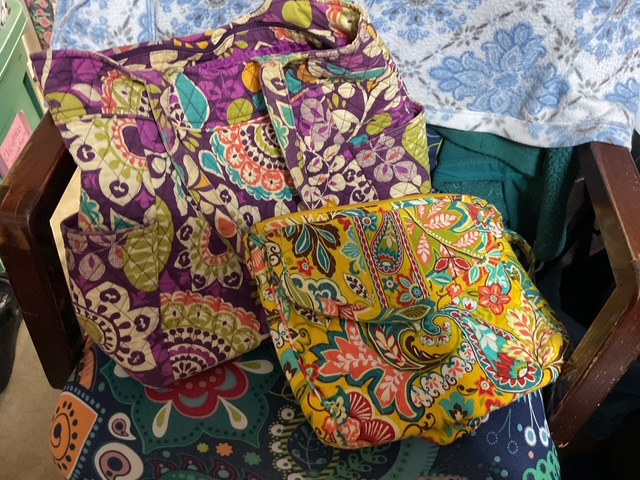 A large, colorful quilted tote bag with purple, green, and floral patterns next to a smaller pouch with vibrant yellow and floral designs, placed on a patterned chair.