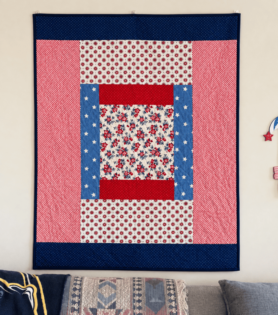 A colorful quilt displayed on a wall, featuring a patchwork design in red, white, and blue fabrics with floral and star patterns.