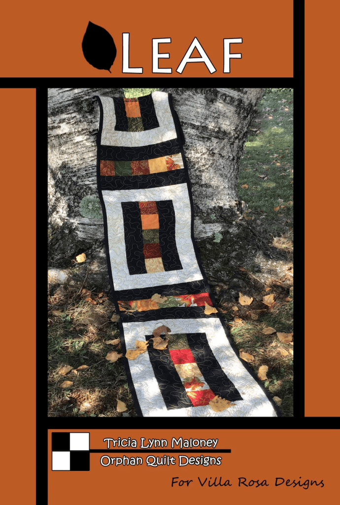 A quilted table runner displayed on grass and autumn leaves, featuring a geometric design with warm colors and black borders. The title 'LEAF' is prominently featured at the top.