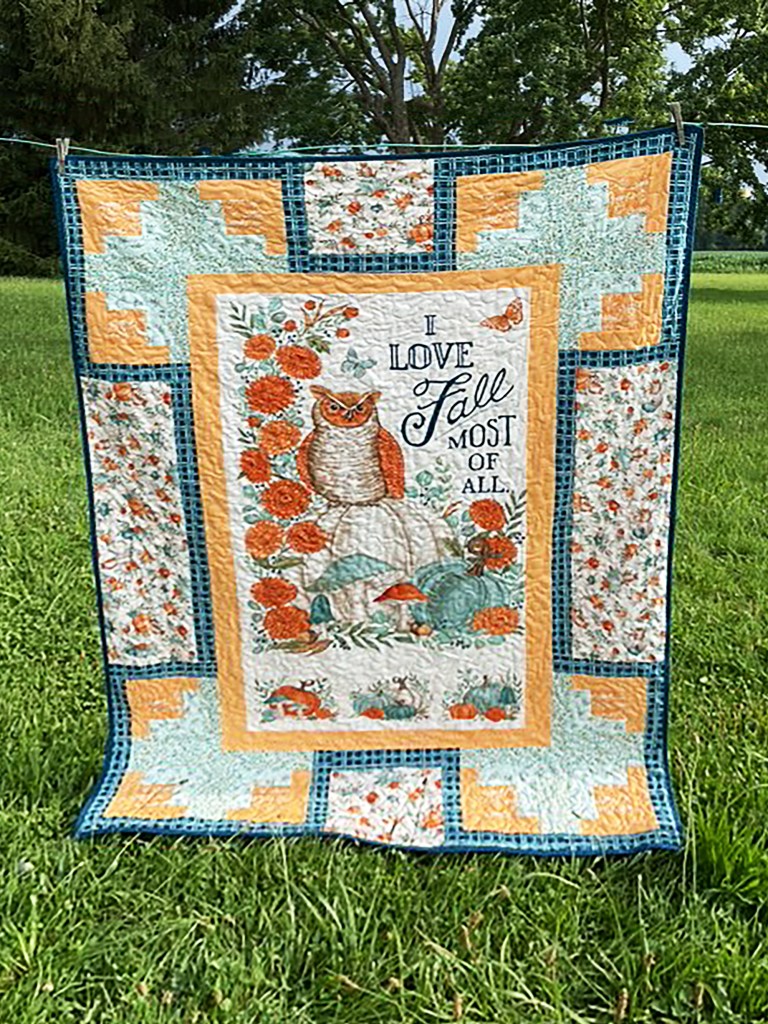 A quilt featuring an owl surrounded by autumn-themed designs, including flowers and mushrooms, with the text 'I love Fall most of all'. The quilt has a colorful patchwork border in shades of orange and blue, displayed on a grassy field.