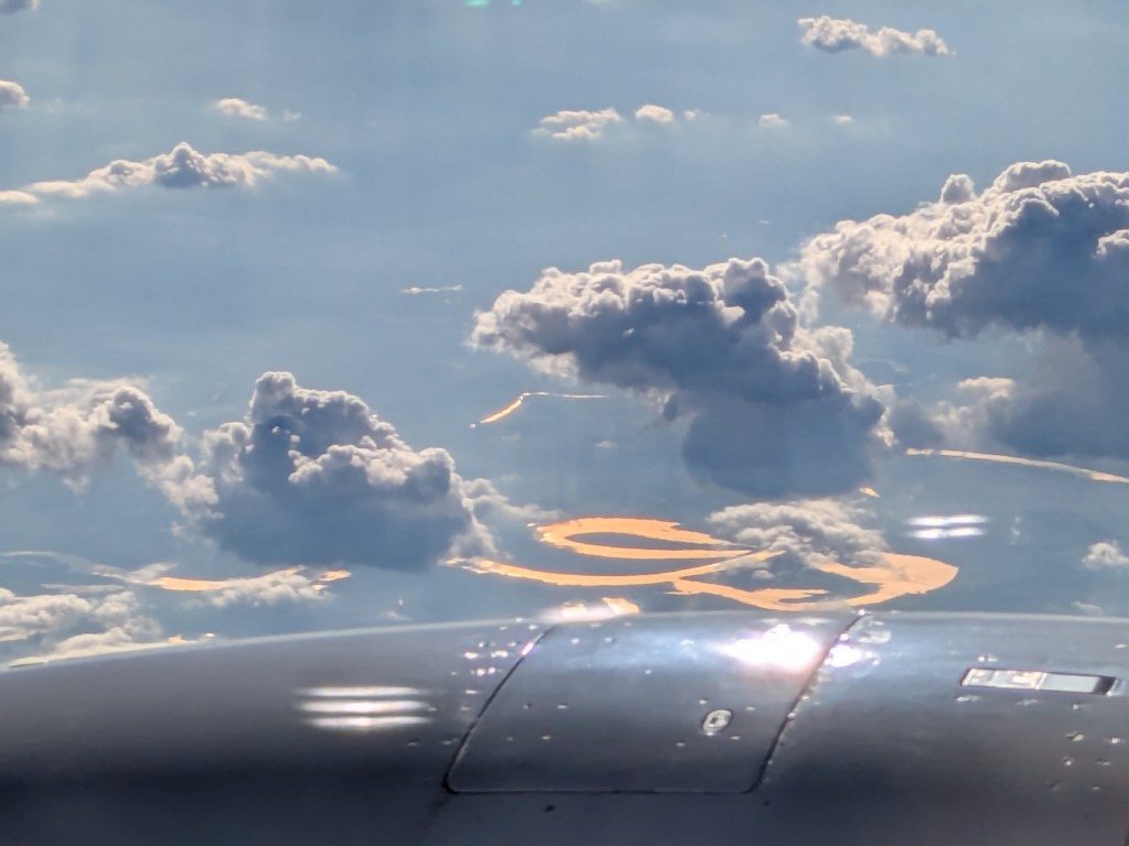 Aerial view of the clouds and a river reflecting sunlight, with part of an airplane wing visible in the foreground.