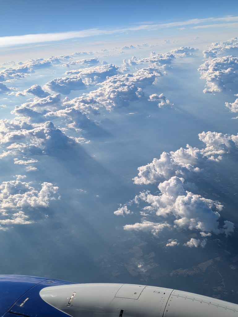 Aerial view of fluffy white clouds under a blue sky with sunlight rays visible. The image shows part of an airplane wing in the lower corner.