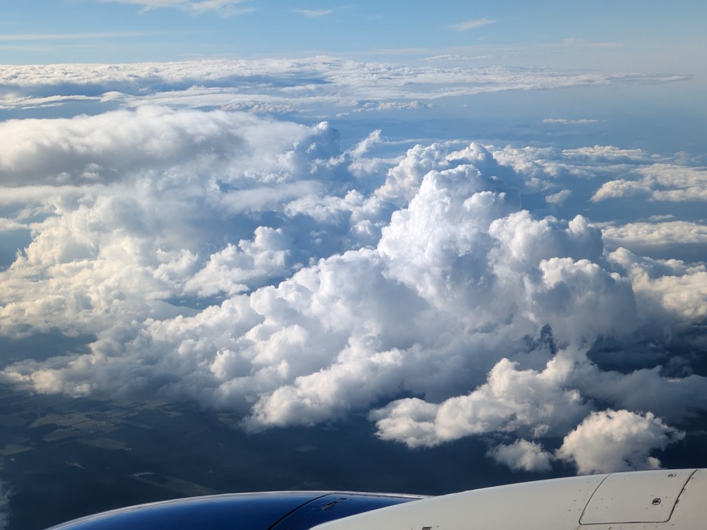 Aerial view of fluffy white clouds against a blue sky, with a partial airplane wing visible in the foreground.