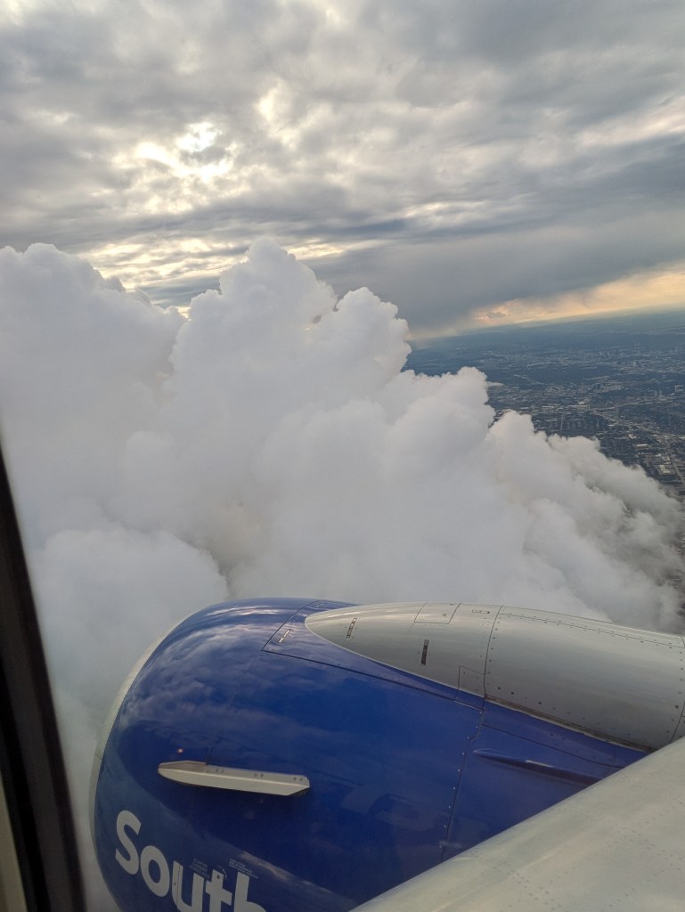 View from an airplane window showing a jet engine and thick white clouds beneath a cloudy sky.