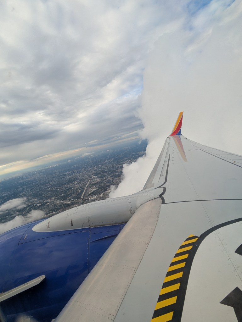 View from an airplane window showing a wing, clouds, and a cityscape below.