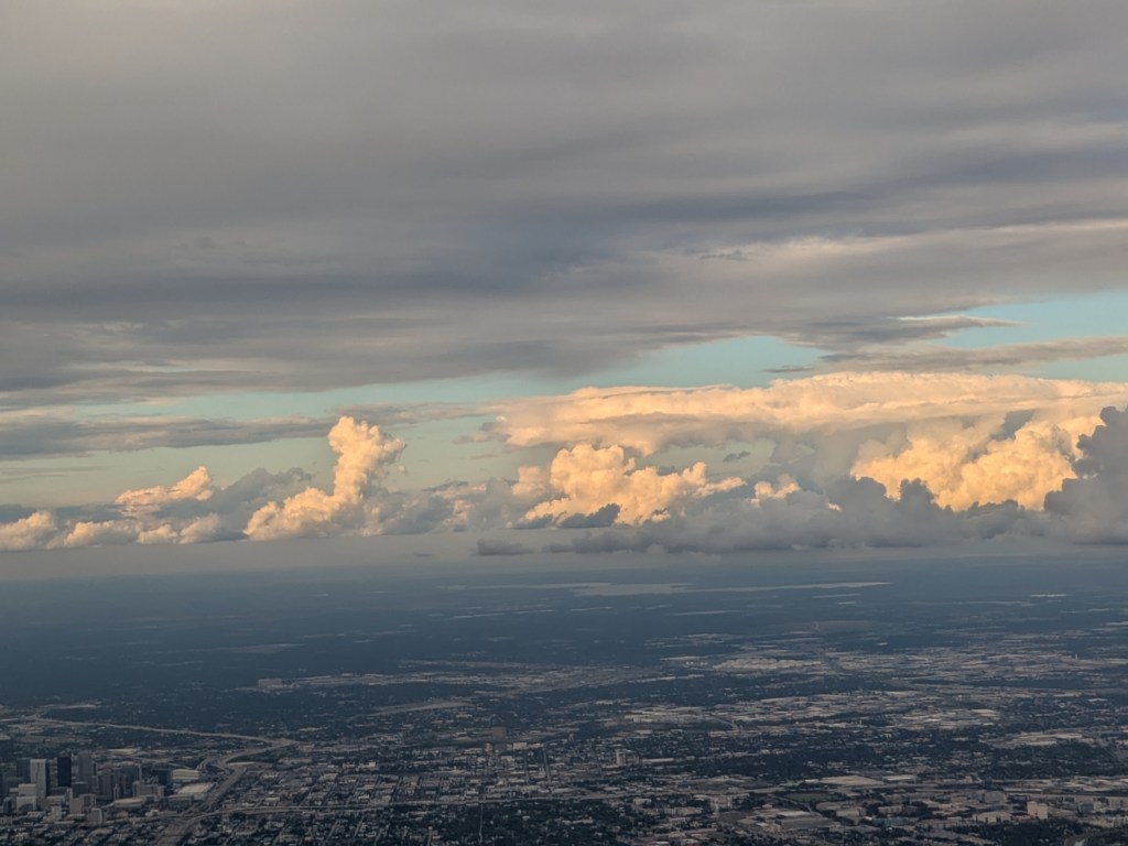 Aerial view of a landscape under a cloudy sky with varying cloud formations and shades of gray and white.