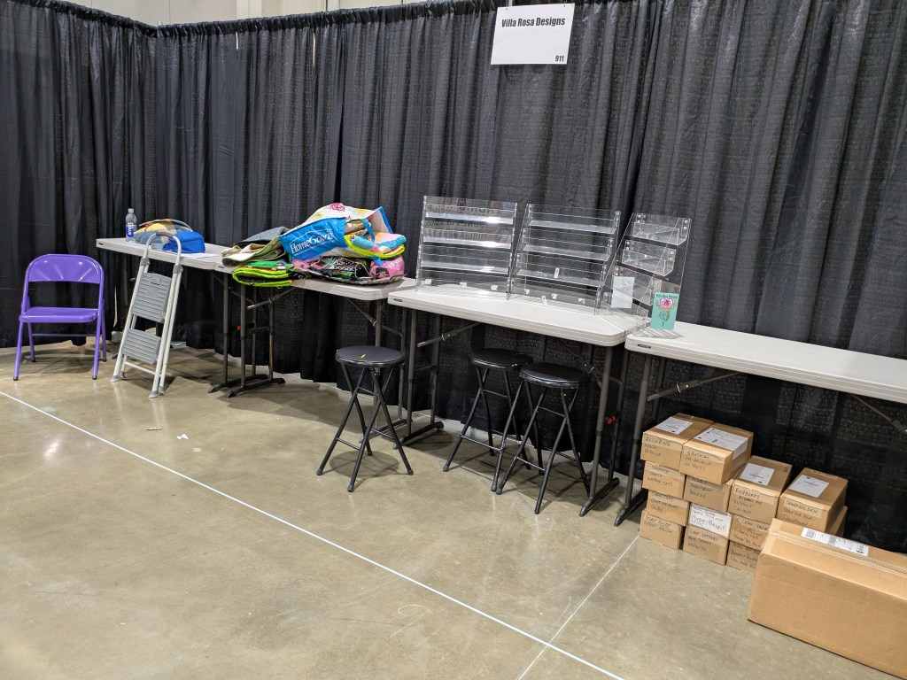 A vendor booth setup displaying tables with various items, including colorful fabric rolls, a water bottle, and packaging boxes. There are empty chairs and a sign indicating the booth name, 'Villa Rosa Designs'. The area is enclosed by a black curtain.
