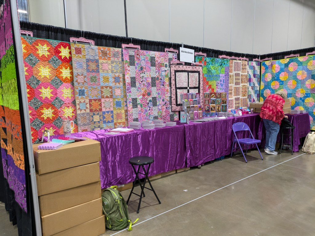 A colorful quilt display at a craft fair booth featuring various intricate quilt patterns and vibrant fabrics, with a vendor organizing items on a table draped in purple fabric.