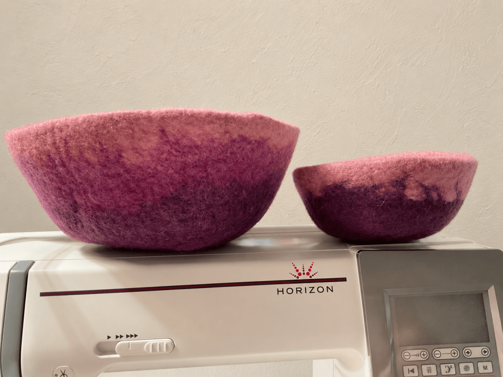Two handmade felt bowls in shades of pink and purple, sitting on top of a sewing machine.