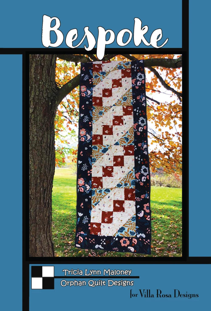 A quilt design titled 'Bespoke' by Tricia Lynn Maloney, hanging from a tree amidst colorful autumn leaves, showcasing a patterned quilt with a floral and geometric design.