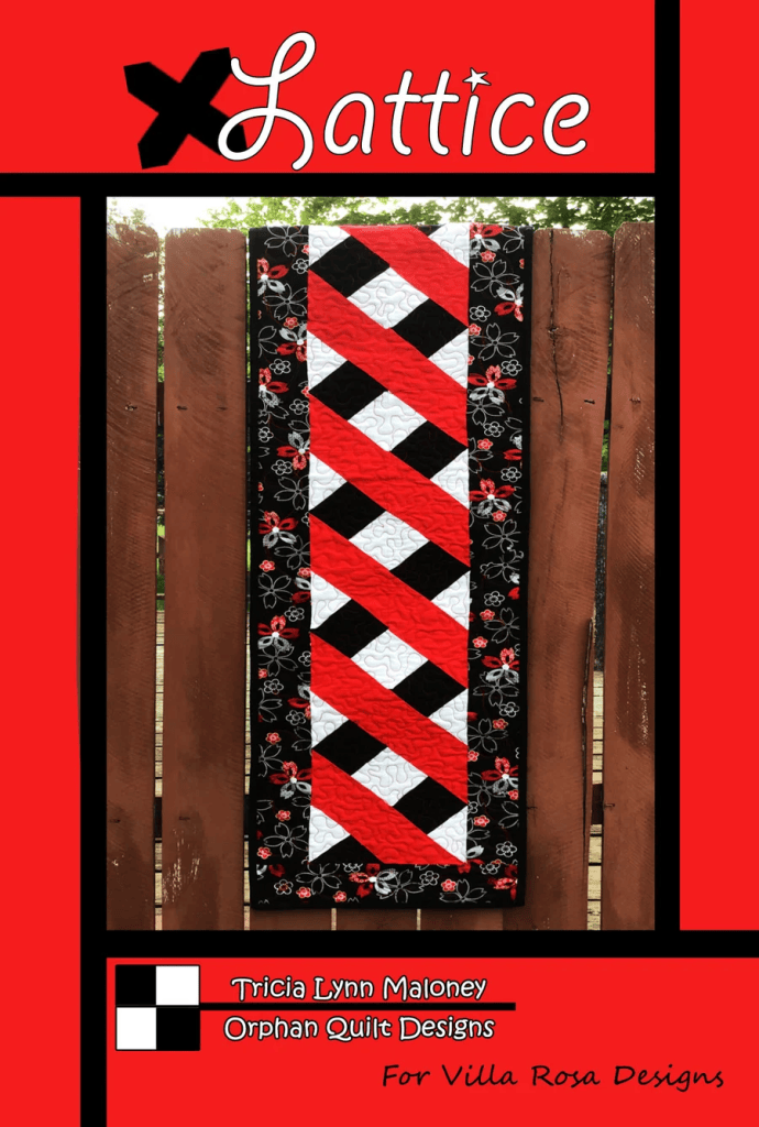 A vibrant quilt designed by Tricia Lynn Maloney titled 'Lattice', featuring a zigzag pattern in black, red, and white with floral accents. The quilt is displayed against a wooden fence.