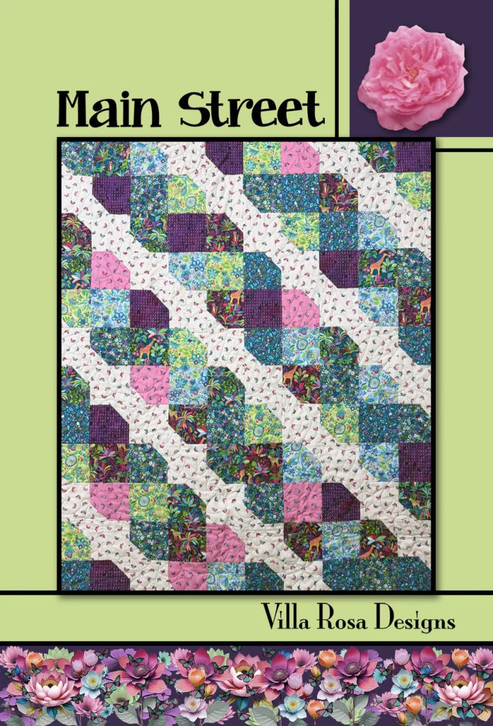 A colorful quilt featuring a geometric pattern in shades of pink, purple, green, and blue, titled 'Main Street' by Villa Rosa Designs, against a light green background.
