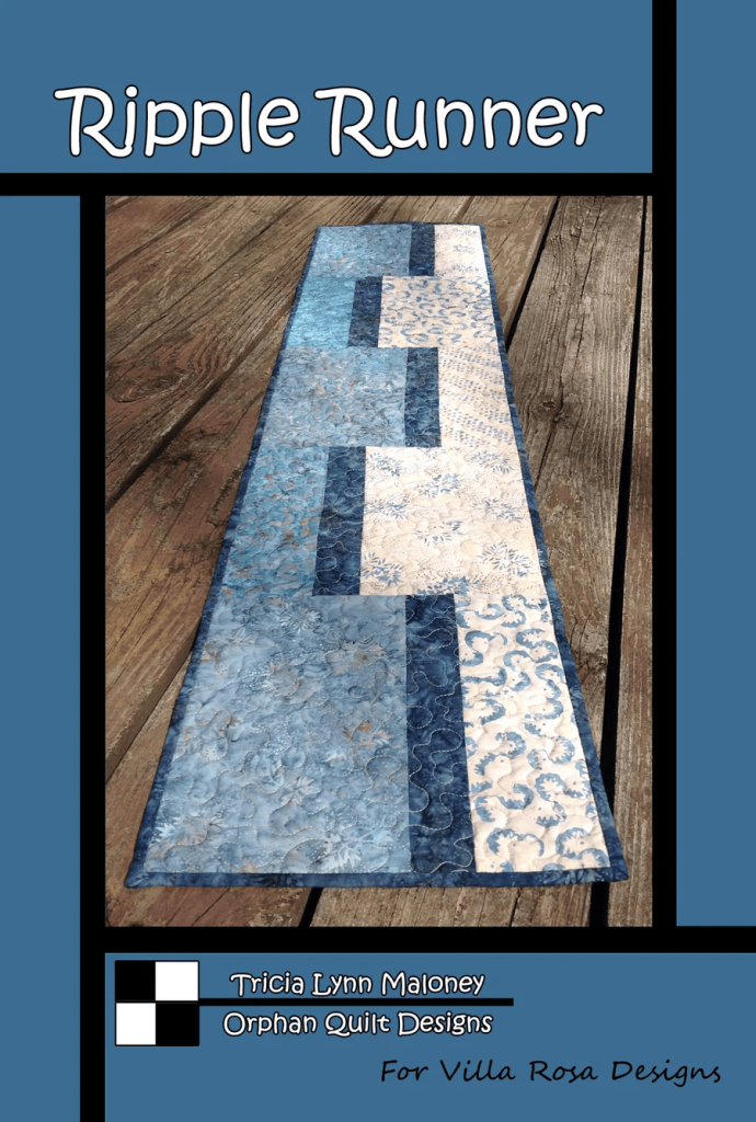 A blue and white quilted table runner called 'Ripple Runner' displayed on a wooden surface, designed by Tricia Lynn Maloney for Orphan Quilt Designs.