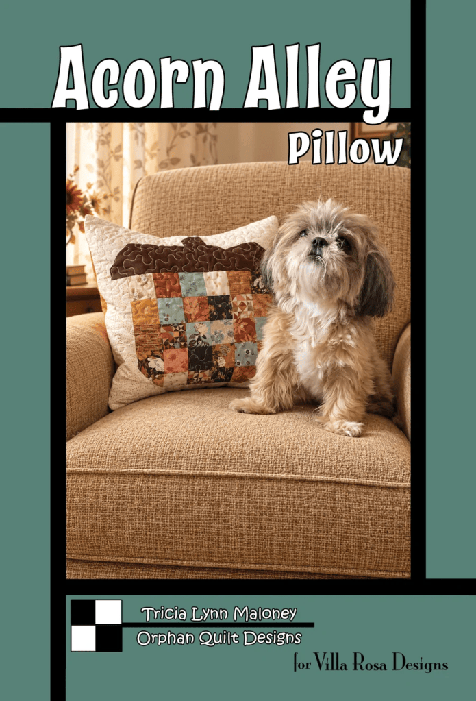 A cozy living room scene featuring a beige sofa with a decorative patchwork pillow titled 'Acorn Alley Pillow' by Tricia Lynn Maloney. A small, fluffy dog sits on the sofa, looking towards the camera.