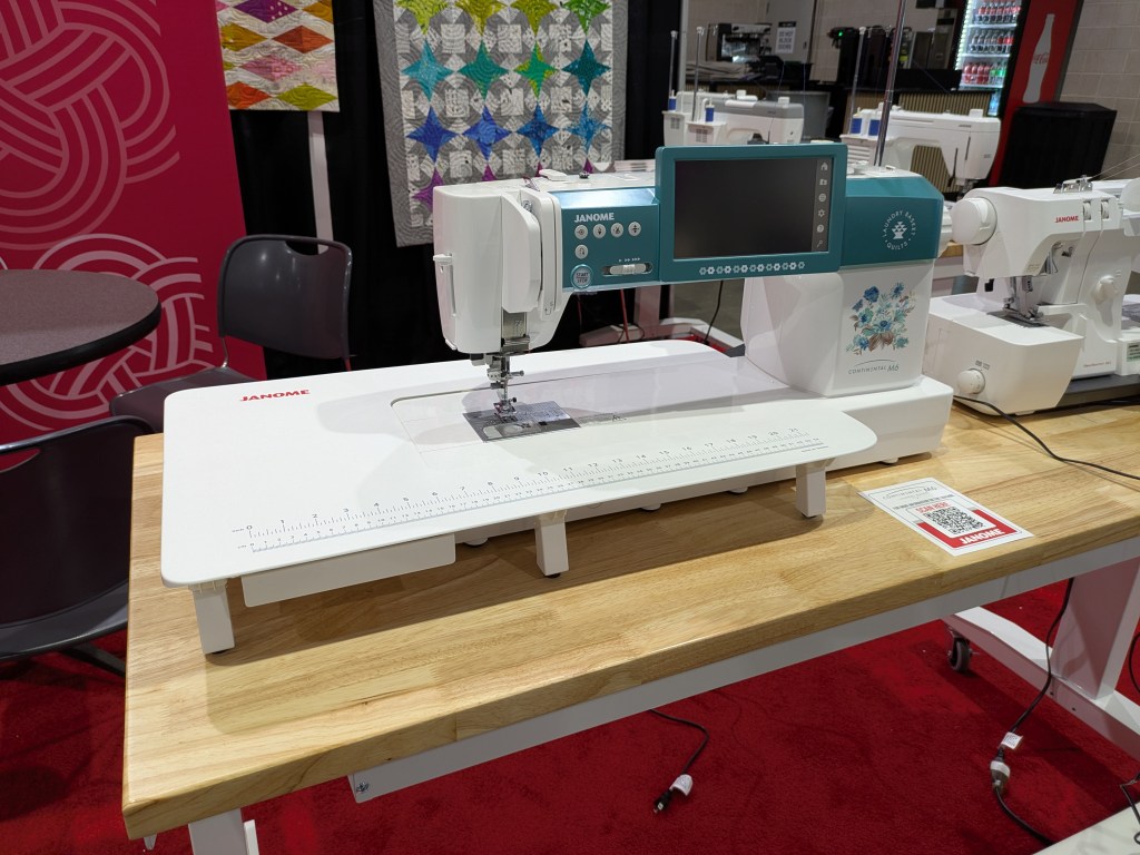 A Janome sewing machine displayed on a wooden table, showcasing a modern design with a blue and white color scheme and a digital interface.