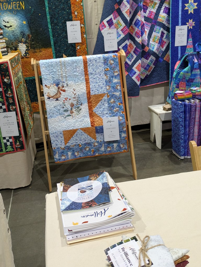 A display of colorful quilts at a craft show, featuring a prominent blue quilt with a star and winter-themed imagery, alongside other quilts and quilting books on a table.
