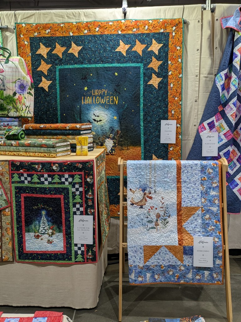 A display of colorful quilts in a fabric shop, featuring a Halloween-themed quilt with a blue background and orange border, and seasonal quilts hanging on the right, along with fabric rolls and decorative items on a table.