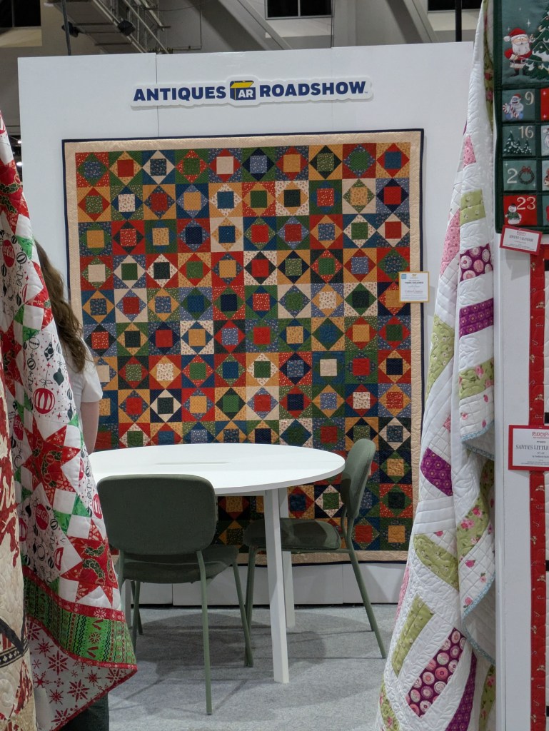 A colorful quilt hanging on a wall at an event, with a round white table and green chairs in front. Two additional quilts are draped on either side, creating a cozy display atmosphere.
