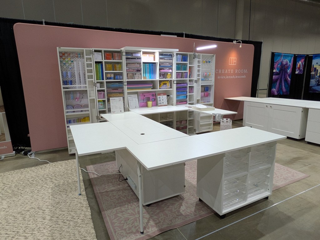 A modern craft workspace featuring a large white table and an organized shelving unit filled with colorful crafting supplies, set against a light pink backdrop.