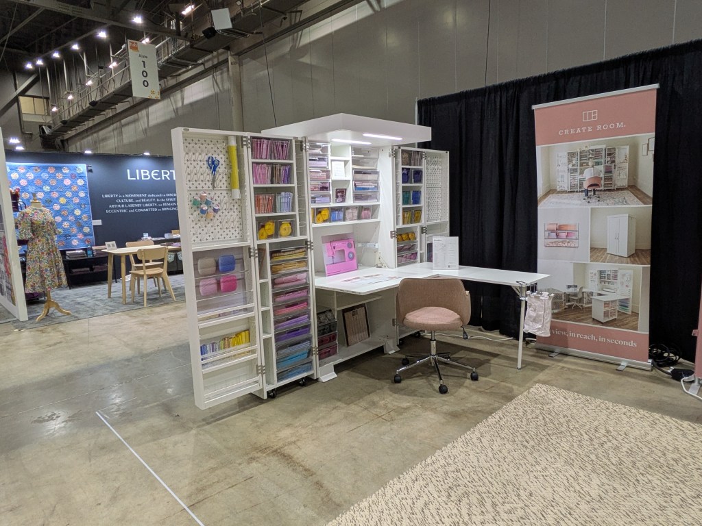 A modern craft room setup featuring a white desk with a pink chair, surrounded by organized storage units filled with crafting materials, paper, and supplies. A promotional banner for 'Create Room' is displayed nearby, suggesting efficient crafting solutions. The background contains a textile display and a small seating area.