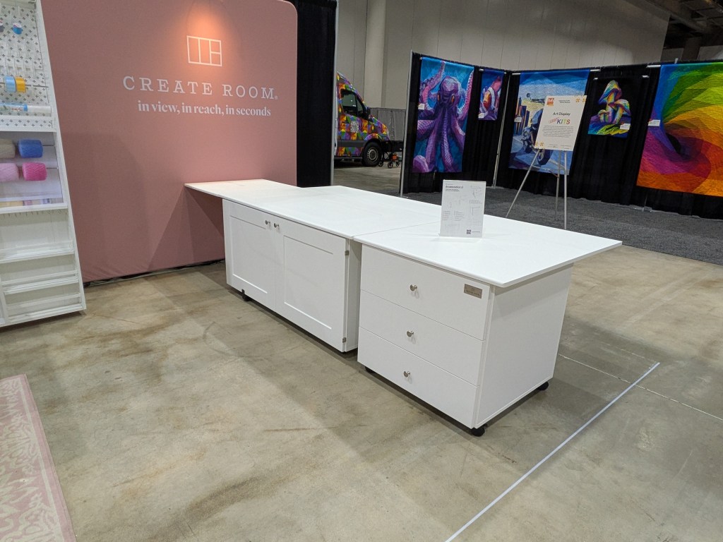 A white craft table with drawers displayed in a trade show setting, featuring a pink backdrop with the text 'CREATE ROOM. in view, in reach, in seconds.'