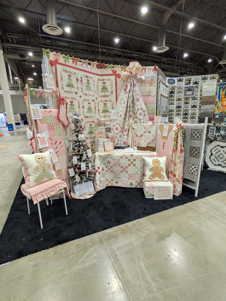 A festive display featuring various holiday-themed quilts, a small decorated Christmas tree, and decorative pillows, set up in a booth with light-colored flooring and a black carpet.