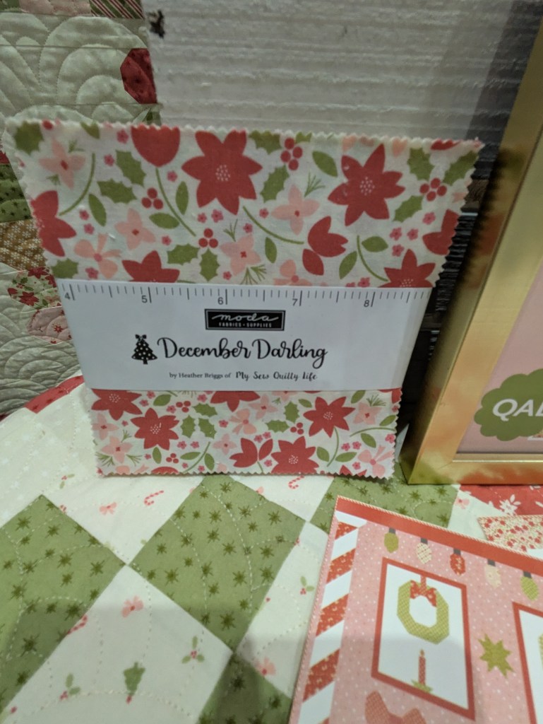 A fabric bundle titled 'December Darling' by Heather Briggs, featuring floral patterns in red, pink, and green. Displayed next to a quilted table and festive cards.