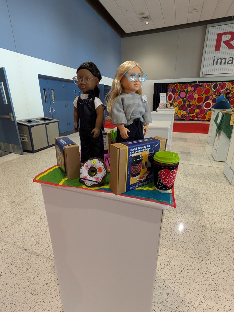 Two dolls displayed on a colorful fabric, surrounded by craft kits and a decorative cup. The left doll has dark hair and glasses, while the right doll has blonde hair and a sweater.