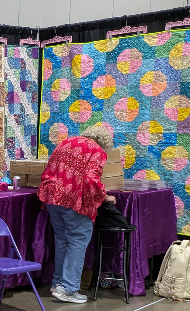 A woman in a red patterned blouse is leaning over a table covered with a purple tablecloth, arranging items. Colorful quilted wall hangings with geometric patterns are visible in the background.