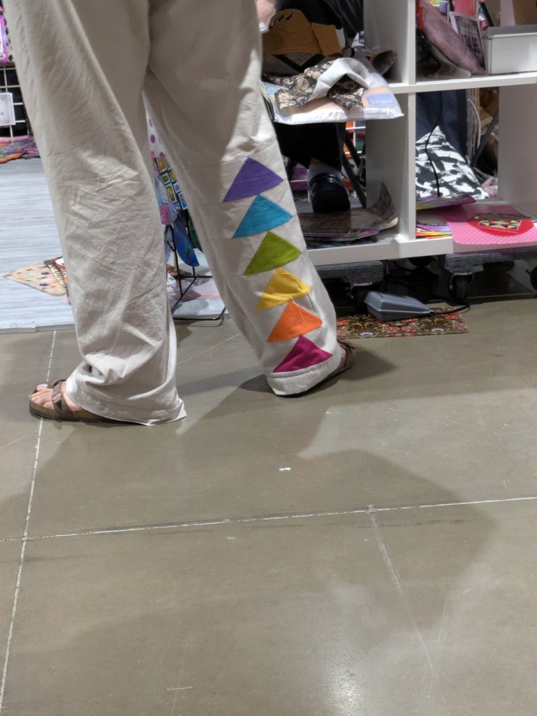 A person wearing beige pants adorned with a colorful triangular pattern in shades of purple, green, yellow, and orange on the side.