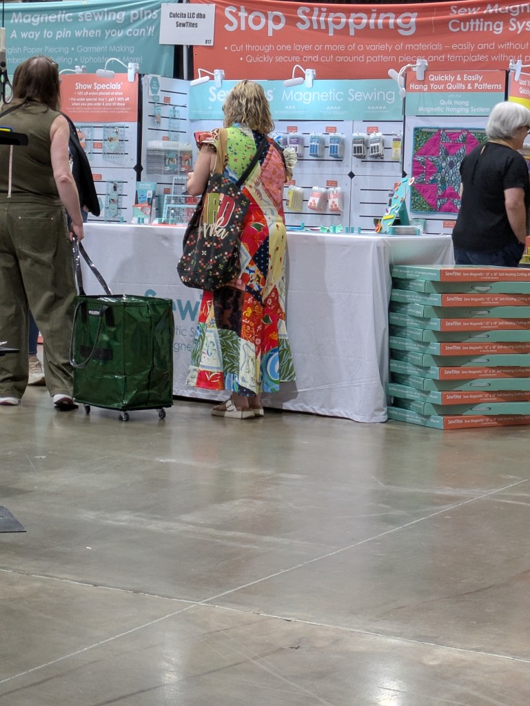 Visitors exploring a sewing booth at a trade show, featuring magnetic sewing tools and supplies. One person carries a green wheeled bag and another wears a colorful dress with floral patterns.