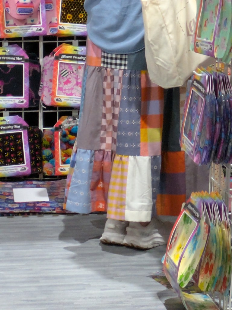 A person standing in a store aisle wearing a colorful patchwork skirt and light-colored shoes, with various craft supplies visible in the background.