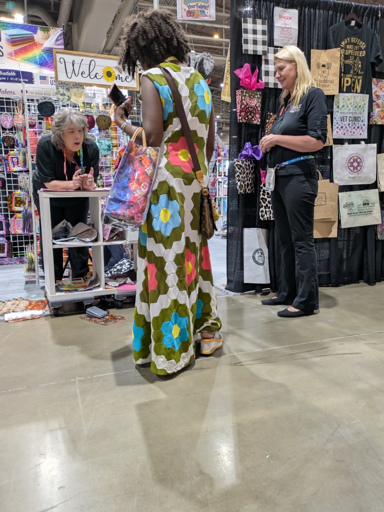 A woman with curly hair wearing a brightly colored floral dress interacts with a vendor at a booth, while another vendor smiles in the background. The booth features various fabric and craft items, along with a welcome sign.
