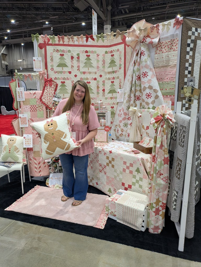 A woman stands in a booth displaying a variety of colorful quilts and fabric, holding a gingerbread-themed pillow. The booth features patterned quilts with Christmas trees, stars, and various festive designs.