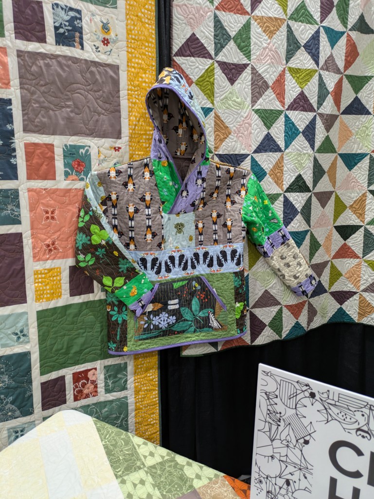 A colorful patchwork quilted jacket with a hood, featuring various patterns including animals and plants, displayed against a backdrop of patterned quilts.