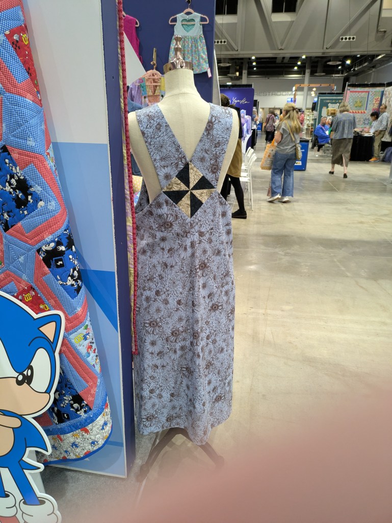 Back view of a floral-patterned dress on a mannequin, featuring a unique cross-back design. The setting includes colorful quilt displays and people browsing at an event.