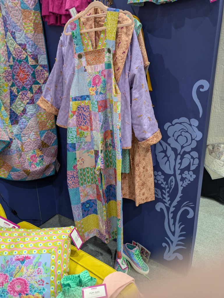 Colorful patchwork overalls hanging on a display, alongside a matching purple jacket and various patterned accessories, against a blue wall.