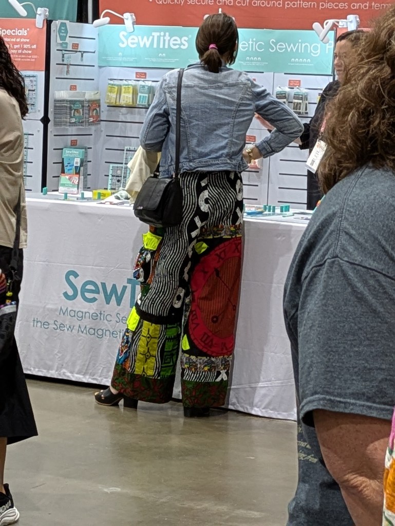 A person wearing colorful, patterned wide-leg pants stands in front of a display table at a sewing convention, with various sewing supplies visible in the background.